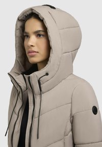 Pufferjacke in hellem Beige mit Kapuze, diagonal gesteppte Muster, Reißverschluss und schwarze Hardware-Details.