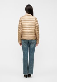 Πανωφόρι τύπου puffer σε ροδακινί χρώμα με οριζόντιο καπιτονέ σχέδιο, στρογγυλεμένο γιακά και ορατές ραφές· συνδυασμένο με μπλε τζιν παντελόνι ίσιας γραμμής.