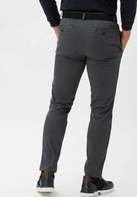 Man wearing slim-fit grey checked trousers, black belt, black long-sleeve shirt, and black sneakers with white soles, hands in pockets.