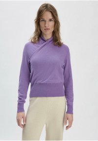 Lila Wickelstyle-Pullover aus weichem, strukturiertem Strick, mit langen Ärmeln und geripptem Saum. Kombiniert mit cremefarbenen, leichten Hosen.