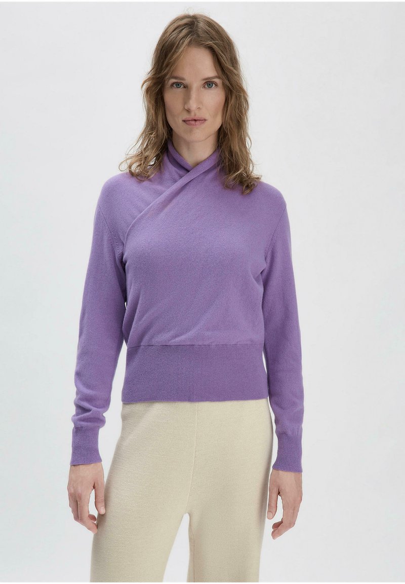 Lila Wickelstyle-Pullover aus weichem, strukturiertem Strick, mit langen Ärmeln und geripptem Saum. Kombiniert mit cremefarbenen, leichten Hosen.