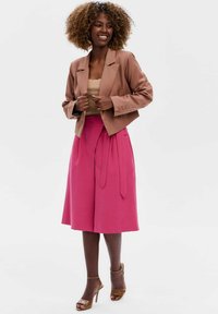 Bruine cropped blazer over een beige top, gecombineerd met een rozekleurige rok tot de knie met een strikceintuur. Bruine sandalen met hakken maken de outfit af.
