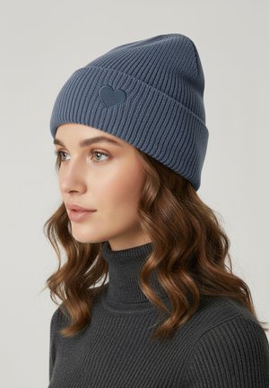 Knit blue beanie with ribbed texture and a heart-shaped accent on the front, worn with a dark turtleneck sweater.