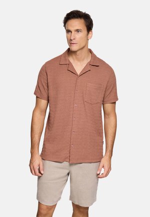 Hombre vistiendo una camisa con botones de manga corta de color óxido texturizada y pantalones cortos beige claro, de pie con expresión neutral frente a un fondo blanco.