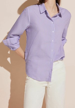 Kobieta ubrana w jasnofioletową i białą koszulę w pionowe paski z długim rękawem podwiniętym do łokci oraz białe spodnie, na beżowym tle.