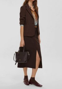 Blazer marrone con texture sopra una camicia marrone a fantasia, abbinato a una gonna marrone con uno spacco laterale. Accessoriato con una borsa marrone scuro e scarpe borgogna.