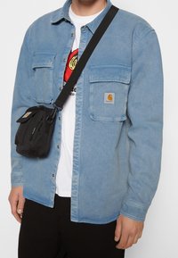 Camicia di jeans azzurro chiaro con due tasche sul petto. Borsa a tracolla nera con un'unica tasca con zip collegata a una tracolla nera.