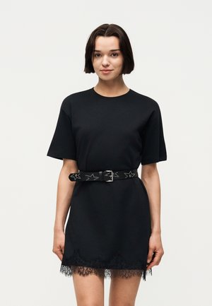 Nuori nainen pukeutuneena mustaan lyhythihainen mekkoon, jossa on pitsireuna ja tähdillä koristeltu niittivyö, seisoo yksivärisen valkoisen taustan edessä.