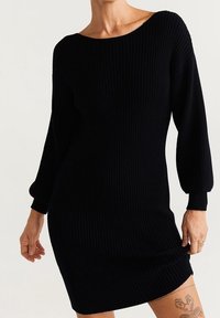 Robe noire en tricot à côtes avec encolure bateau, manches longues bouffantes et silhouette cintrée. Texture douce, ourlet à mi-genoux.
