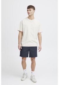 Lichtbeige katoenen T-shirt met korte mouwen, gecombineerd met donkerblauwe korte broek, gedragen met witte sneakers en sokken. Eenvoudig, casual ontwerp.