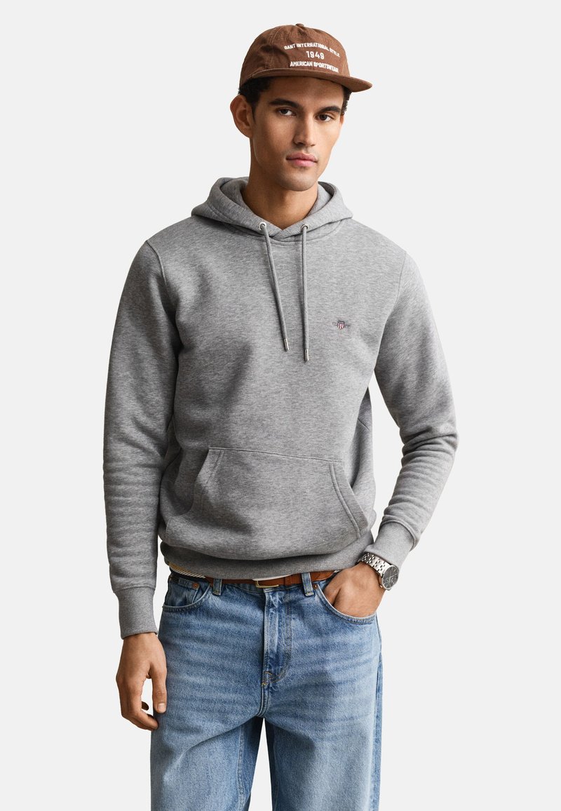 Hombre que lleva una gorra marrón, sudadera con capucha gris con bolsillo frontal, vaqueros azules y un reloj de pulsera, de pie con una mano en el bolsillo.