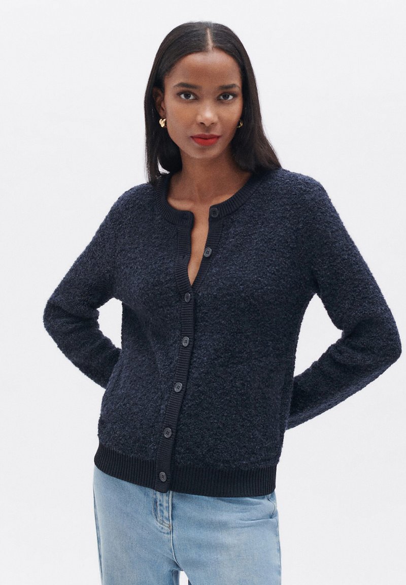 Cardigan blu navy in tessuto strutturato con scollatura rotonda e chiusura a bottoni, featuring polsini e orlo a coste. Vestibilità standard, tessuto morbido.