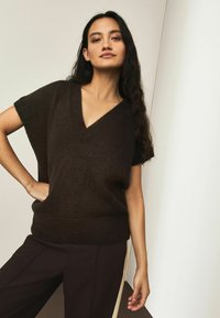 V-hals kortærmet sweater i mørkebrunt, lavet af blødt, struktureret stof. Har en afslappet pasform og ribbet kantdetalje. Kombineret med mørke bukser.