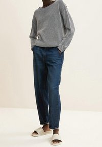 Helles graues Strickpullover mit weitem Ausschnitt, kombiniert mit dunkelblauen Jeans. Abgerundet mit beigen, gesteppten Slide-Sandalen.