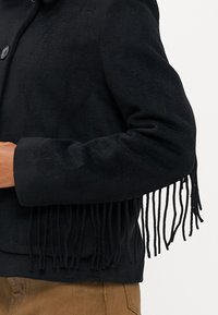 Černá zkrácená bunda vyrobená z měkké látky s třásněmi na rukávech. Obsahuje zapínání na knoflík a zaoblený límec.