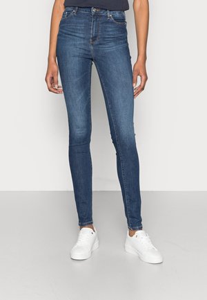 Personne portant un jean skinny bleu et des baskets blanches, debout devant un fond clair uni.