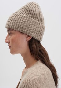 Vrouw met lang bruin haar, draagt een beige geribbelde gebreide muts en een bijpassende beige trui, staande naar links tegen een effen lichte achtergrond.