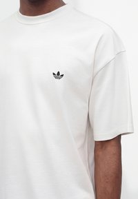 Gros plan d'une personne portant un t-shirt blanc uni avec un petit logo noir Adidas trefoil sur la poitrine gauche.
