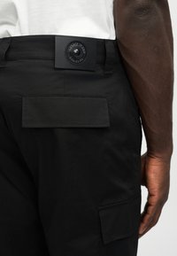 Μαύρο παντελόνι με τσέπες με καπάκι και δερμάτινο λογότυπο Versace Jeans Couture στη μέση, φορεμένο από ένα άτομο με λευκό πουκάμισο.