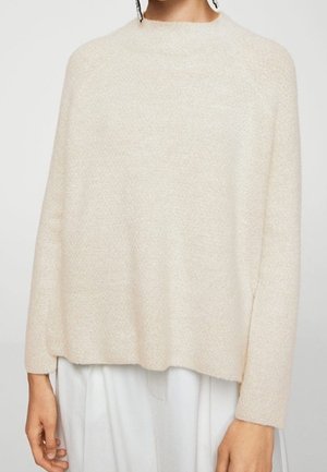 Vrouw draagt een losse, lange mouwen, crèmekleurige, zachte trui gecombineerd met witte broek tegen een effen achtergrond.