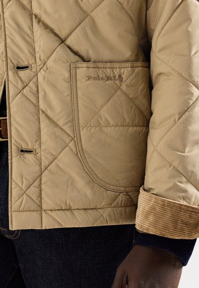 Beżowa pikowana kurtka z dużą boczną kieszenią, mankietem z sztruksu i prążkowanym wykończeniem. Materiał ma gładką fakturę i błyszczące wykończenie.