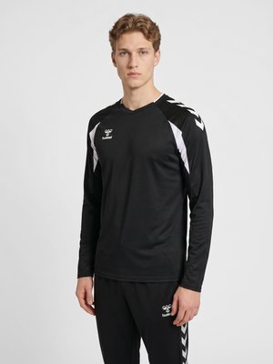 Jonge man draagt een zwart-witte hummel sporttrui met lange mouwen en zwarte hummel broek tegen een effen lichte achtergrond.