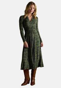 Kobieta w zielonej, wzorzystej sukience midi z długim rękawem, w połączeniu z brązowymi skórzanymi botkami sięgającymi do połowy łydki, stojąca na tle jednolitego tła.