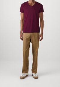 Burgundské bavlněné tričko s výstřihem do v písmene V, kombinované se béžovými kalhotami a bílými teniskami. Tričko má krátké rukávy a slim fit.