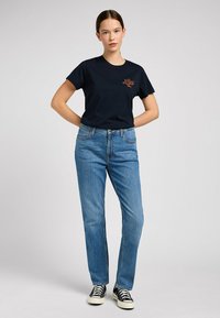Granatowa koszulka z logo "Lee" w kolorze czerwonym, zestawiona z jasnoniebieskimi prostymi dżinsami i czarnymi sneakersami. Prosty, casualowy design, wykonana z bawełny.