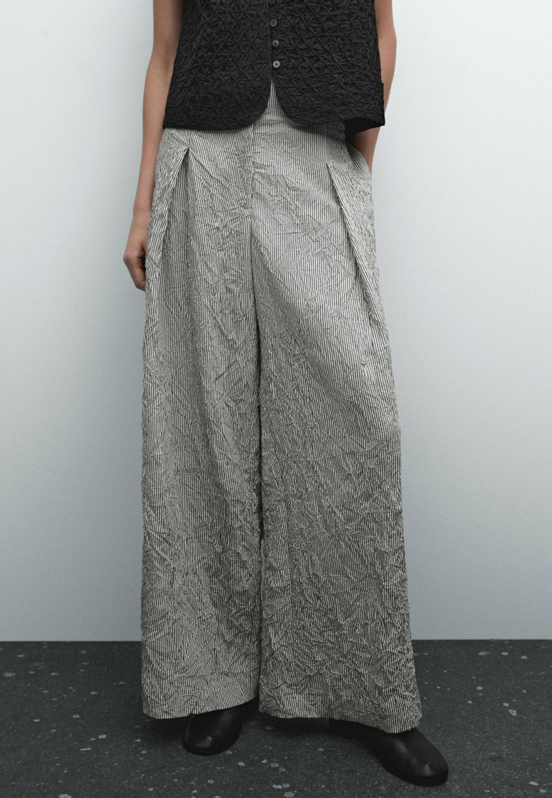 Personne portant un pantalon taille haute, large, froissé de couleur gris clair avec de fines rayures verticales et un gilet noir sans manches texturé, debout sur un sol foncé.
