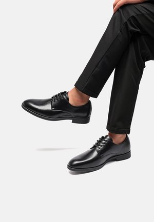Scarpe da uomo in pelle nera con finitura liscia, punta arrotondata e design con lacci. Abbinate a pantaloni neri con risvolto, che mettono in risalto i dettagli delle scarpe.