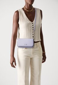 Light purple crossbody bag with smooth texture, rectangular shape, and chain strap. Worn with a cream knitted top and light beige pants.