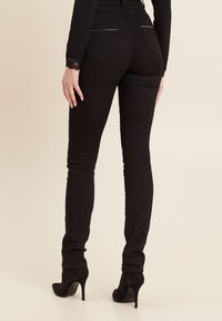 Schwarze Skinny-Jeans mit glatter Textur, ausgestattet mit Gesäßtaschen und silbernen Akzenten. Getragen mit schwarzen High-Heels.