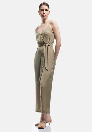 Vrouw staat in een beige mouwloze jumpsuit met zijstrik, draagt beige sandalen met lage hakken tegen een effen witte achtergrond.