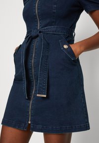 Vestido de mezclilla en azul oscuro con cierre frontal, mangas cortas, cinturón y dos bolsillos con acentos de hardware decorativos.