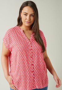 Blusa de patrón de chevron en rojo y crema con cuello esculpido, mangas cortas y cierre frontal con botones, hecha de una tela ligera.