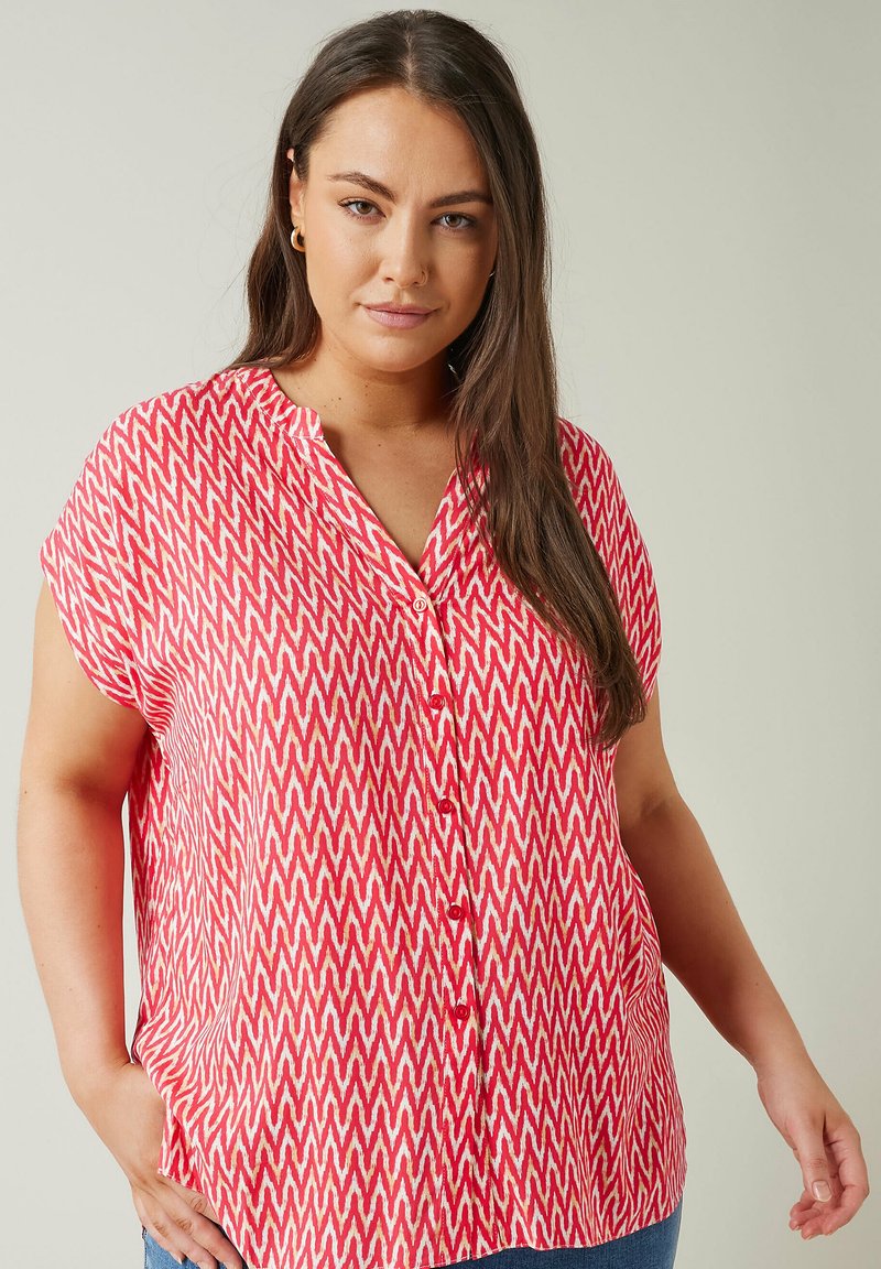 Blusa de patrón de chevron en rojo y crema con cuello esculpido, mangas cortas y cierre frontal con botones, hecha de una tela ligera.