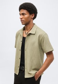 Νεαρός άνδρας με κοντά σγουρά μαλλιά, φοράει ανοιχτοπράσινο πουκάμισο με κοντά μανίκια και κουμπιά, μαύρο αμάνικο μπλουζάκι και κολιέ, με το χέρι στην τσέπη, κοιτάζοντας προς τα αριστερά.