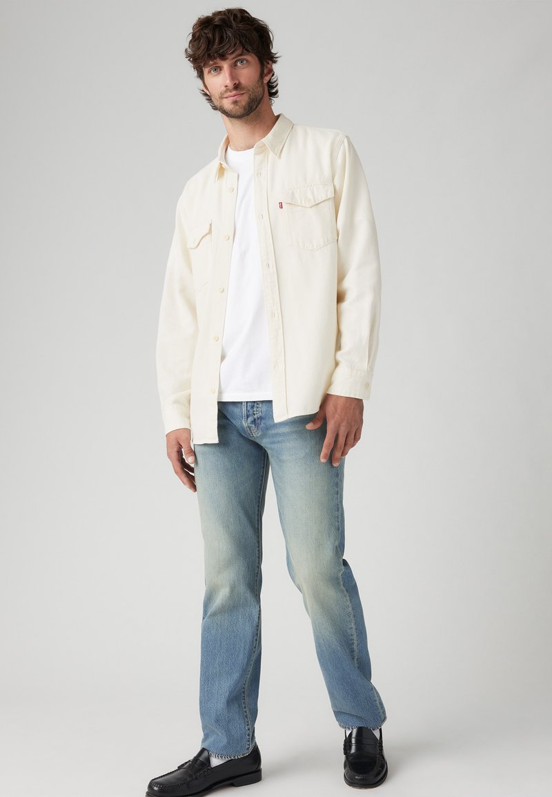 Chemise crème à boutons sur un t-shirt blanc, associée à un jean bleu clair et des mocassins noirs. Détails notables : poches poitrine et coupe décontractée.