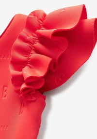 Czerwony materiał z falbanami o teksturowanej powierzchni, z wyraźnym detalem falbany. Materiał wydaje się gładki, z widocznym wytłoczonym logo.