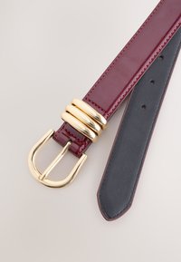 Burgunderroter Lackledergürtel mit einer goldfarbenen Doppeldornschnalle, die drei zylindrische Akzente aufweist, und einer glatten schwarzen Rückseite.