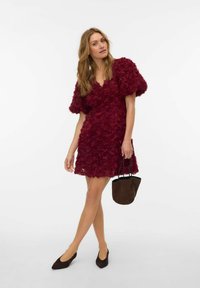 Bordowa sukienka pokryta kwiatowymi aplikacjami z bufkami, zestawiona z ciemnobrązowymi butami i małą brązową torebką. Gładka faktura.