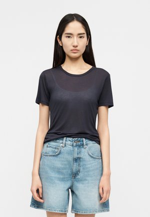 Νεαρή γυναίκα με μακριά σκούρα μαλλιά, ντυμένη με διαφανές μαύρο κοντομάνικο τοπ και ανοιχτά μπλε τζιν σορτς, που στέκεται μπροστά σε απλό φόντο.