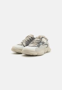 Sneakers gjorda av mesh och syntetiska material, med en färgskala i beige, grått och blått, samt en kraftig sula och vågiga mönster.