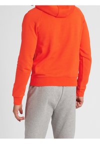 Sudadera naranja con capucha de textura suave, con dobladillo y puños acanalados. Combinada con pantalones de chándal gris claro.