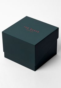 Tamsiai žalia kartoninė dėžutė su blizgiu paviršiumi. Raudonas "TED BAKER LONDON" tekstas viršuje. Kvadratinė forma su atverčiamu dangteliu.