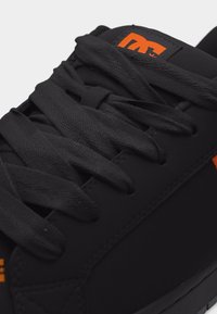 Μαύρο αθλητικό παπούτσι με χοντρές μαύρες κορδόνια, με πορτοκαλί λογότυπο στη γλώσσα και την πλευρά, πάνω σε απλό ανοιχτό φόντο.