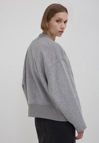 Eine Frau mit kurzen Haaren, die einen lockeren grauen Pullover und schwarze Hosen trägt, schaut über ihre Schulter vor einem einfarbigen hellen Hintergrund.