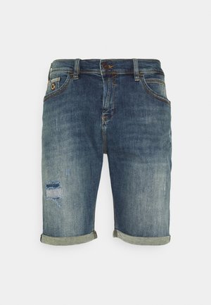 Pantalones cortos de mezclilla azul para hombre con puños enrollados, bolsillos frontales, cierre de botón y un pequeño parche desgastado en la pierna izquierda.