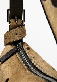 Brauner Felltaschenriemen mit einer dunklen Lederakzent und silbernem Reißverschluss. Das Fell zeigt Flecken und eine glatte Textur, die die Designdetails hervorhebt.